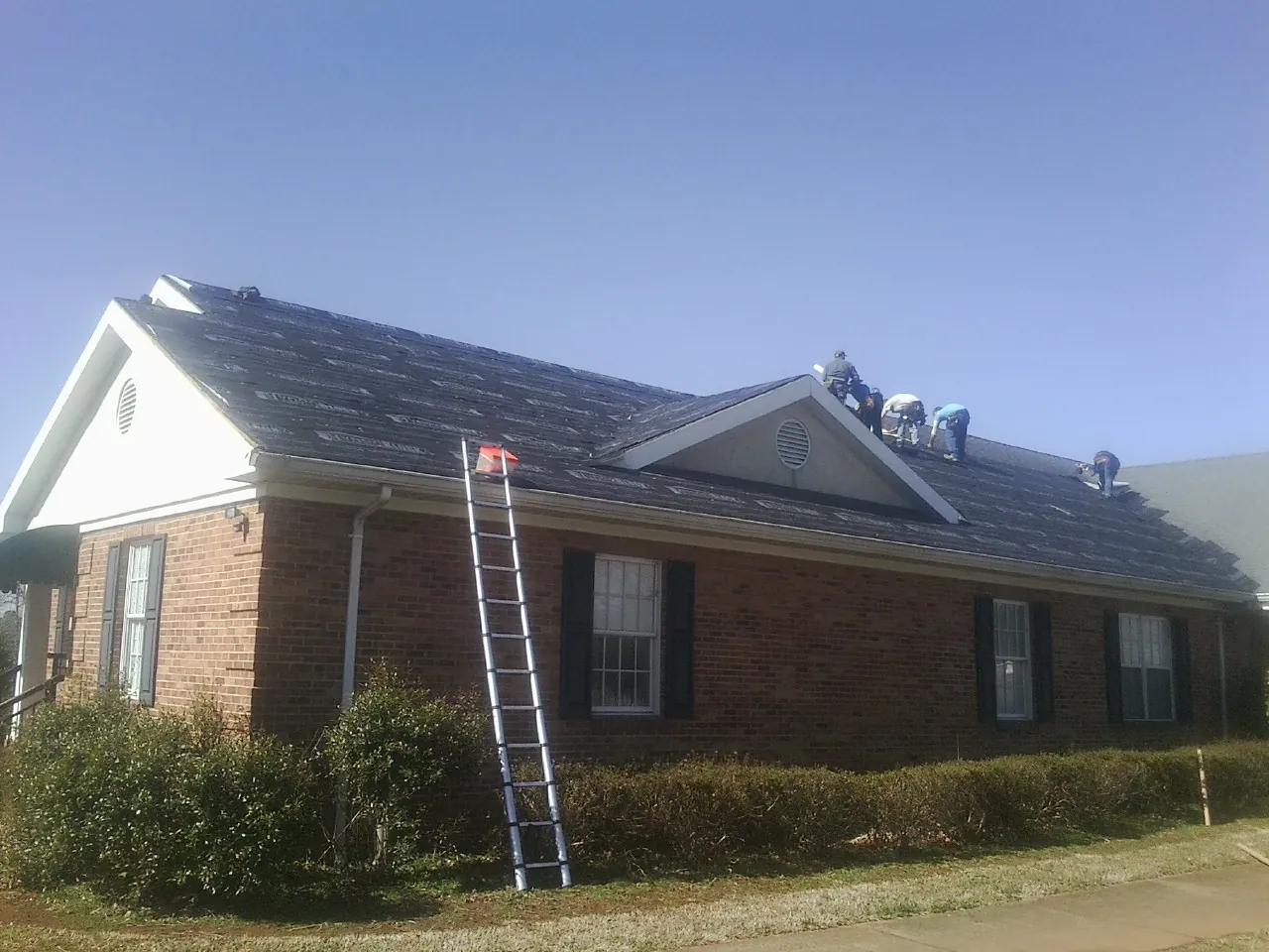 Crew tearing shingles off a brick ranch roof with a ladder set — Metal Roof Repair work by our crew in Rockwell